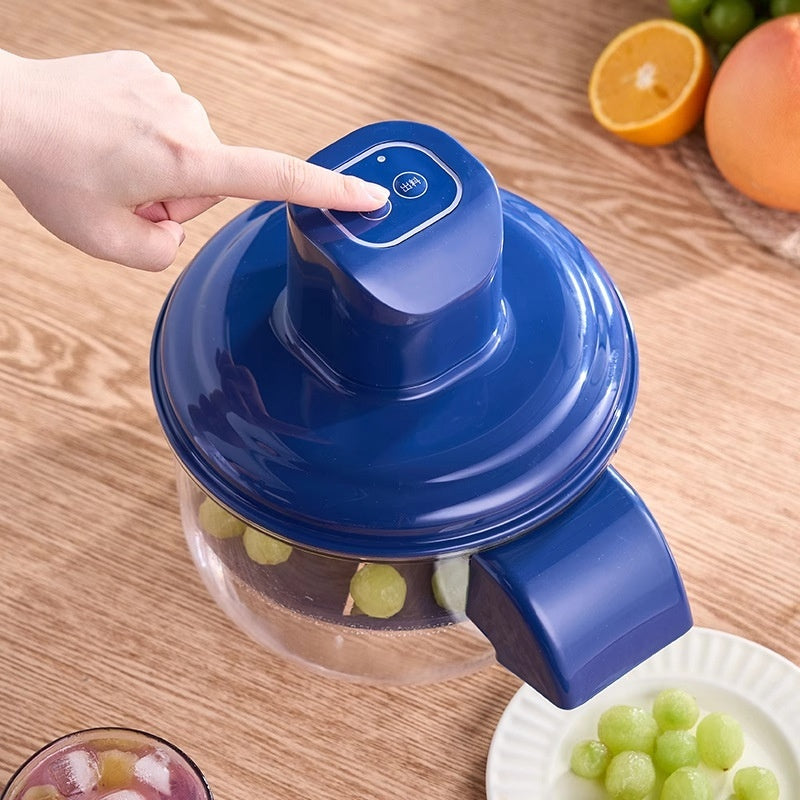 Blue manual juicer with a hand pressing down on a wooden table, surrounded by fruits.
